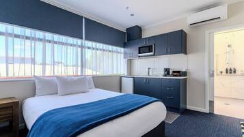 Habitación de hotel con cama, kitchenette pequeña, zona de ducha y ventana con persianas.