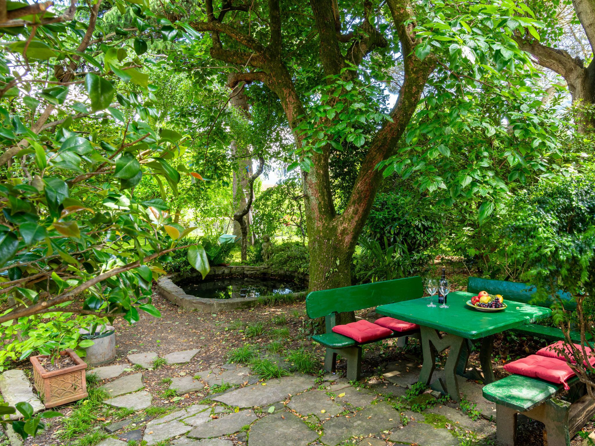 Un jardín con un sendero de piedra, un banco verde con cojines rojos, y una mesa con una bandeja de frutas y vasos.