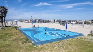 An outdoor basketball court with a blue surface, surrounded by a grassy area and a beach in the background.
