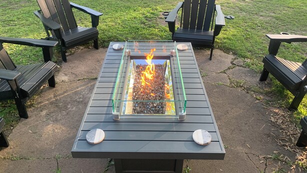 A fire pit table with four Adirondack chairs around it, set on a concrete patio with a grassy field in the background.