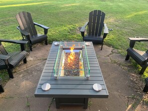 A fire pit table with four Adirondack chairs around it, set on a concrete patio with a grassy field in the background. -  Golf Getaway Sleeps 8 • King Bed • Arcade • Bumper Pool • Full Kitchen (Farmington)