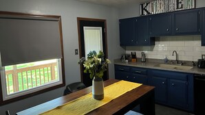 A kitchen with blue cabinets, a wooden table with a yellow placemat, and a window with a view of a wooden deck.