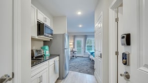 A modern kitchen with white cabinetry, a microwave, a toaster, and a coffee maker. There is a dining area with a table and chairs, and a view of a balcony with a railing. - No Cleaning Fee! Brand New Double Queen Studio Condo w/ Waterfront Pool + Beach (Southport)