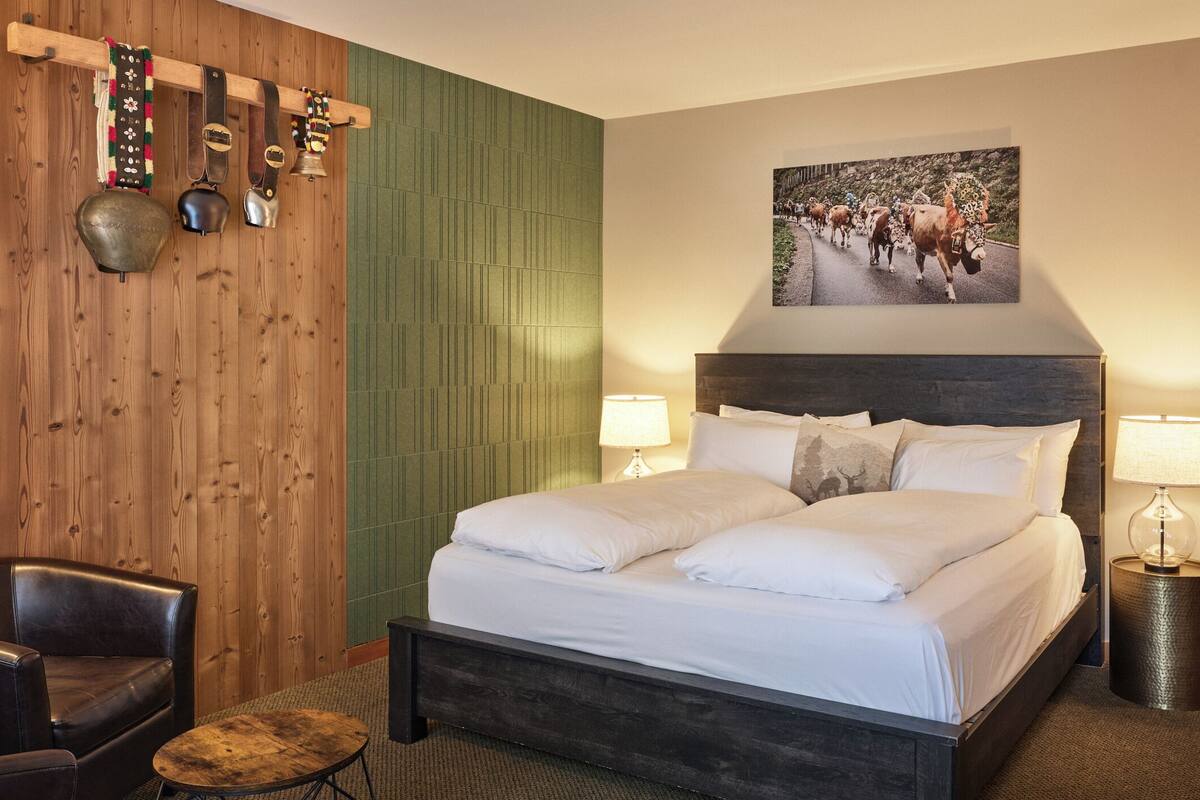 Une chambre d’hôtel avec une tête de lit en bois, un lit avec du linge de lit blanc, deux lampes de chevet, une chaise et une horloge murale.
