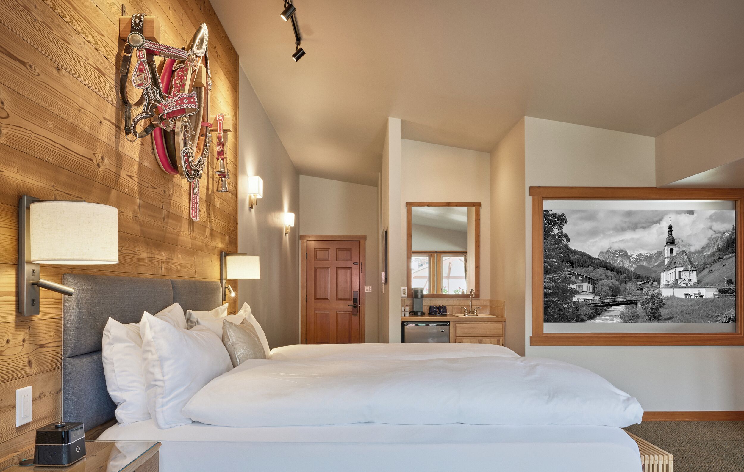 Una habitación de hotel con cabecera de madera, una cama grande, una pared de madera decorada con equipo de equitación, un baño con lavamanos y espejo, y un cuadro paisajístico blanco y negro en la pared.