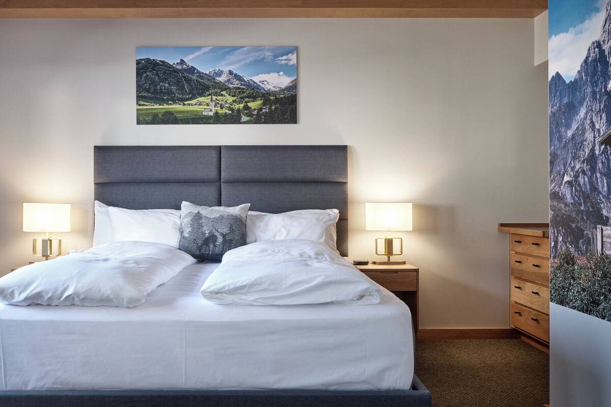 Un lit bien fait, avec du linge de lit blanc et deux oreillers, flanqué de deux tables de chevet sur lesquelles reposent des lampes, dans une pièce où un tableau représentant un paysage de montagne est accroché au-dessus du lit et où un grand tableau mural orne le mur de droite.