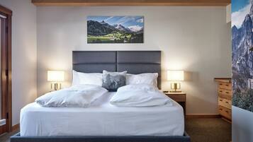 A neatly made bed with white linens and two pillows, flanked by bedside tables with lamps, in a room with a mountain landscape painting above the bed and a large wall painting on the right.