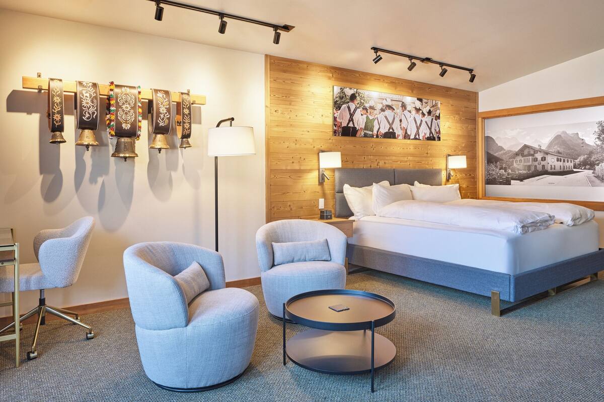 Une chambre d’hôtel comprenant un lit, deux fauteuils, une petite table ronde, un bureau avec une chaise et une lampe fixée au mur.