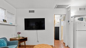 A modern living room with a flat-screen TV mounted on the wall, a wooden side table, a blue armchair, a round ottoman, and a white refrigerator. - Sunlit Palm Cottage in Uptown Phoenix (Phoenix)