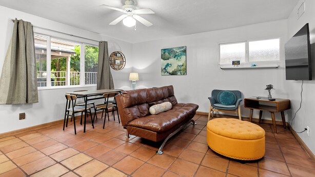 A living room with a brown leather sofa, a yellow ottoman, a dining table with chairs, a ceiling fan, and a TV mounted on the wall. - Sunlit Palm Cottage in Uptown Phoenix (Phoenix)