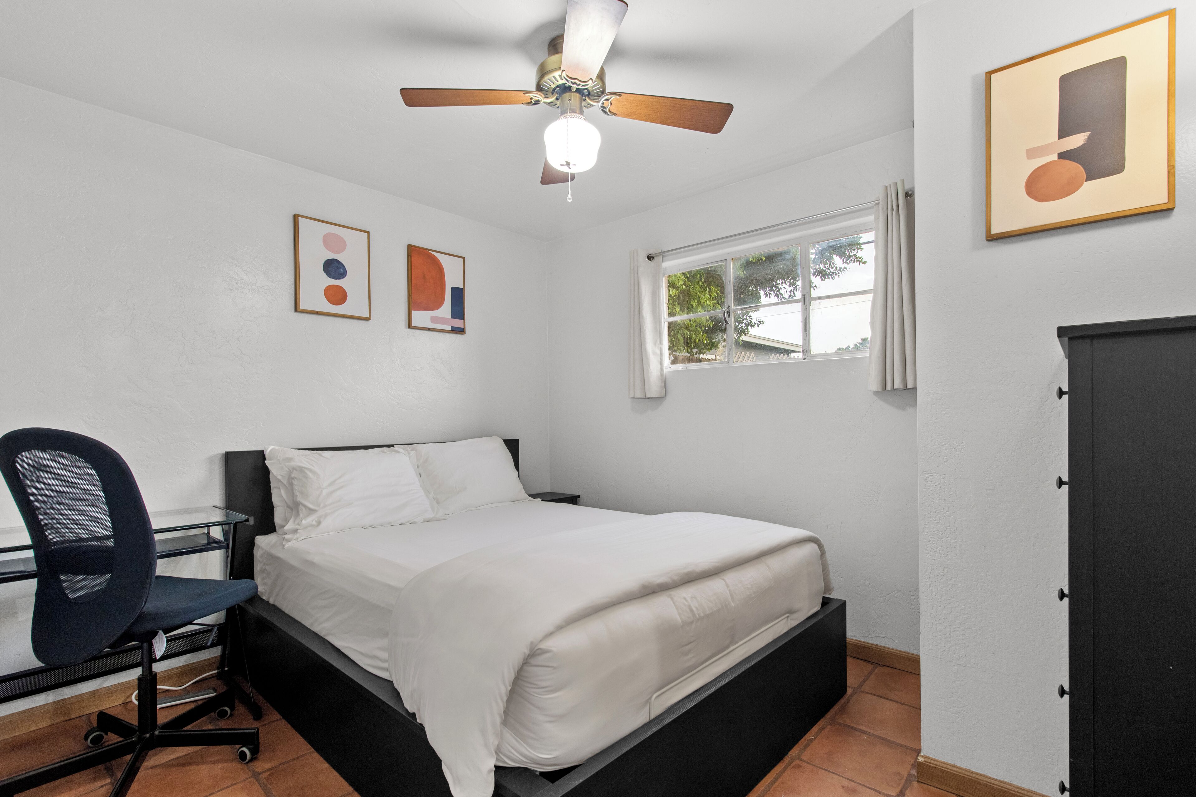 A bedroom with a bed, desk, chair, ceiling fan, window, and artwork on the walls.