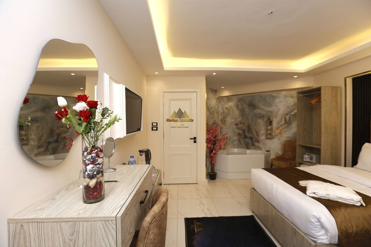 A modern hotel room with a bed, a sitting area with a mirror, a vanity with a vase of flowers, a bathtub, and a large mirror on the wall.
