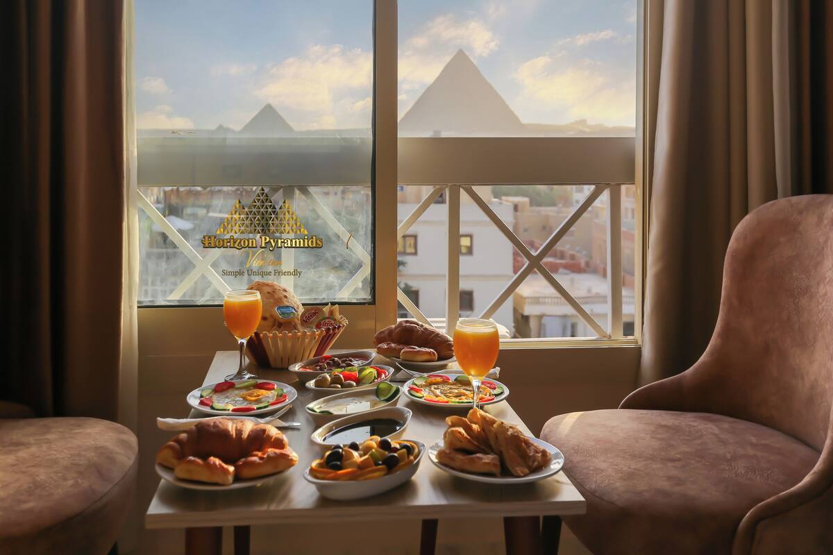 A breakfast table with various dishes and drinks, a view of pyramids through a window, and a chair.