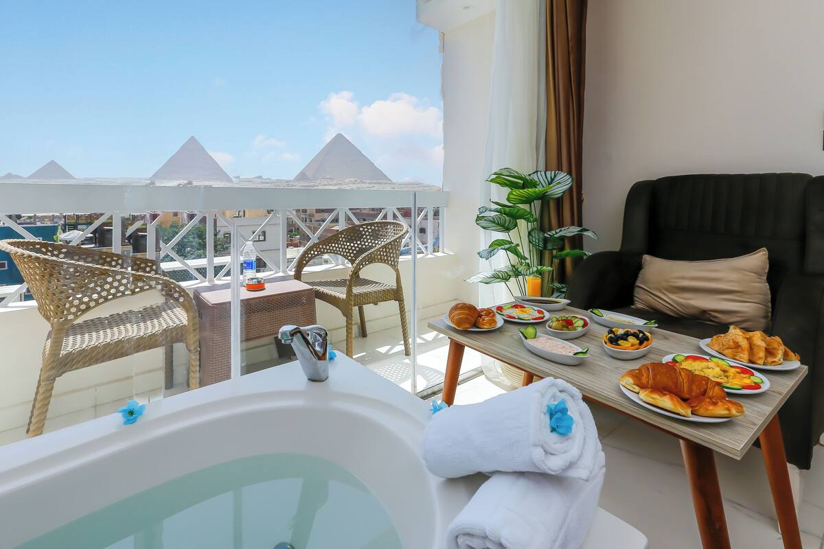 A balcony with a view of pyramids, a wicker chair, a black sofa, a table with breakfast, and a bathtub.