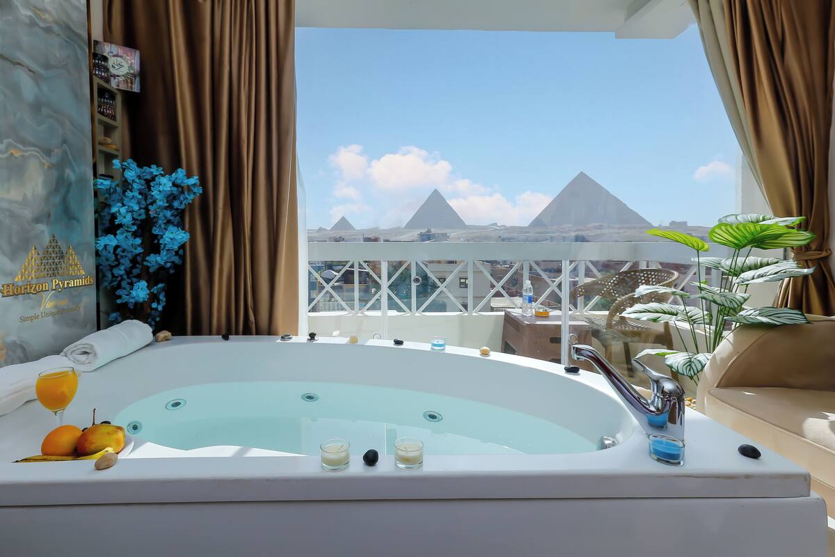A hotel room with a bathtub, a view of the pyramids, and a balcony with plants.