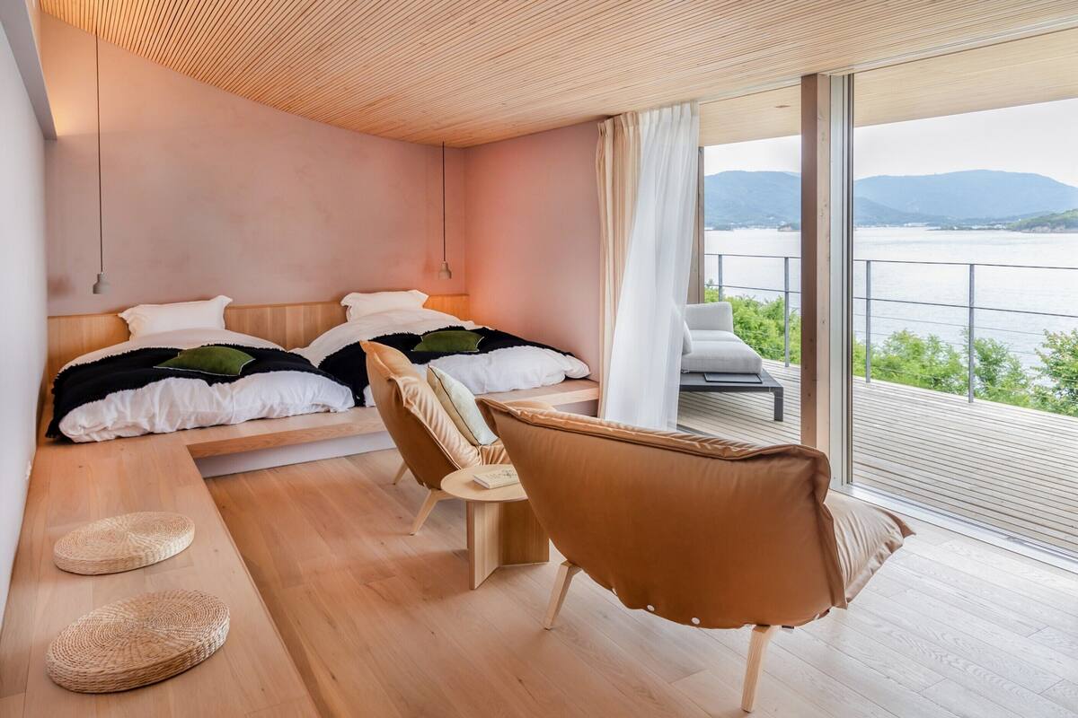 Quarto moderno com uma cama, duas cadeiras, uma mesinha e vista para a água.