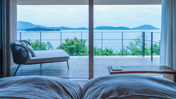 Un dormitorio con un ventanal grande, una cama, una silla y vistas a un cuerpo de agua y montañas.