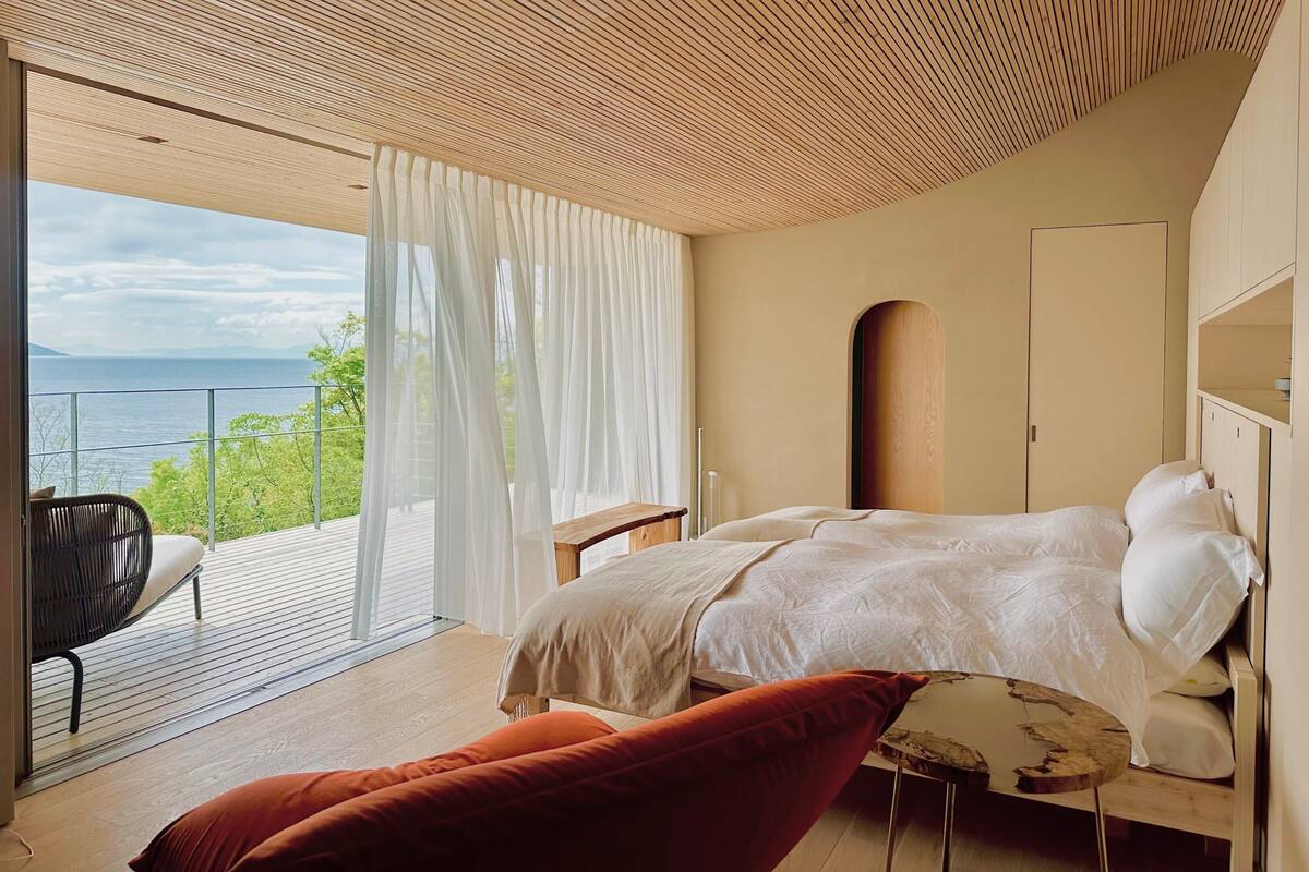 Quarto com cama, uma cadeira, uma mesa e vista para o mar.