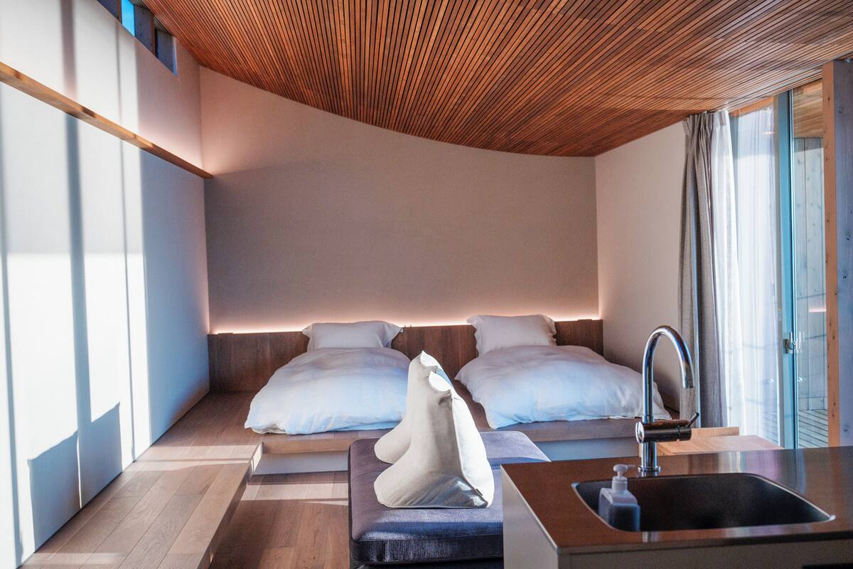 Quarto de hotel moderno com teto de madeira, uma janela grande, cama com roupa branca, mesa de cabeceira e pia.