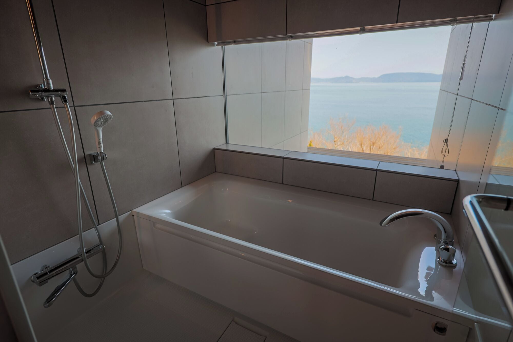 A modern bathroom with a bathtub, a shower, and a window offering a view of the sea.