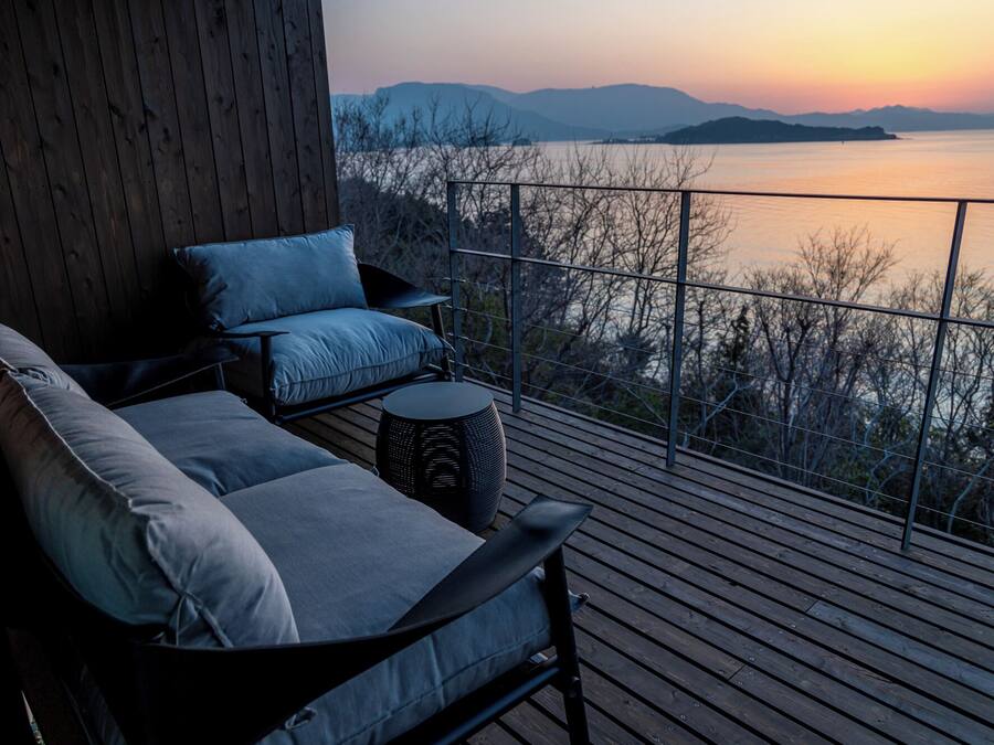A balcony with a sofa and a round table, overlooking a body of water and distant mountains.