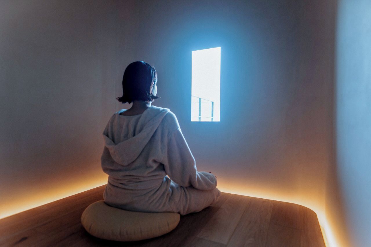 A person sitting on a wooden bench in a room with a window.