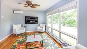Una habitación con ventilador de techo, televisor de pantalla plana, dos sillones blancos, alfombra floral y ventanas amplias con vistas a vegetación.