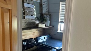 A laundry room with a washing machine and dryer, wooden shelves with storage boxes, and a window with blinds. - THE SPOT, EAST SHORE (Westwood)