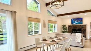 A modern living room with a dining area, large windows, a fireplace, and a TV mounted on the wall. - Fully new 3BR House in heart of Pocono Mountains, Fire Pit! Close to Water Park! (Tobyhanna)