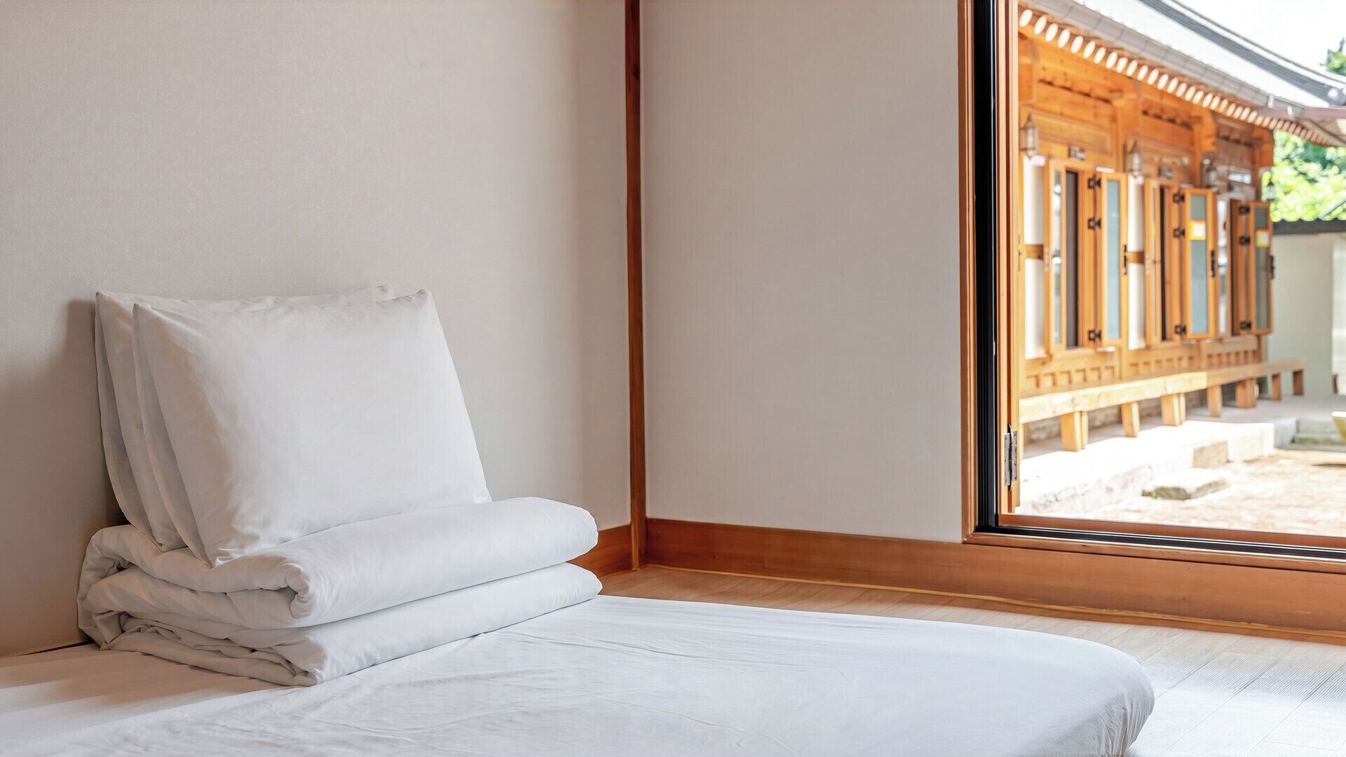 A neatly made bed with white linens in a room with wooden flooring and a large window.