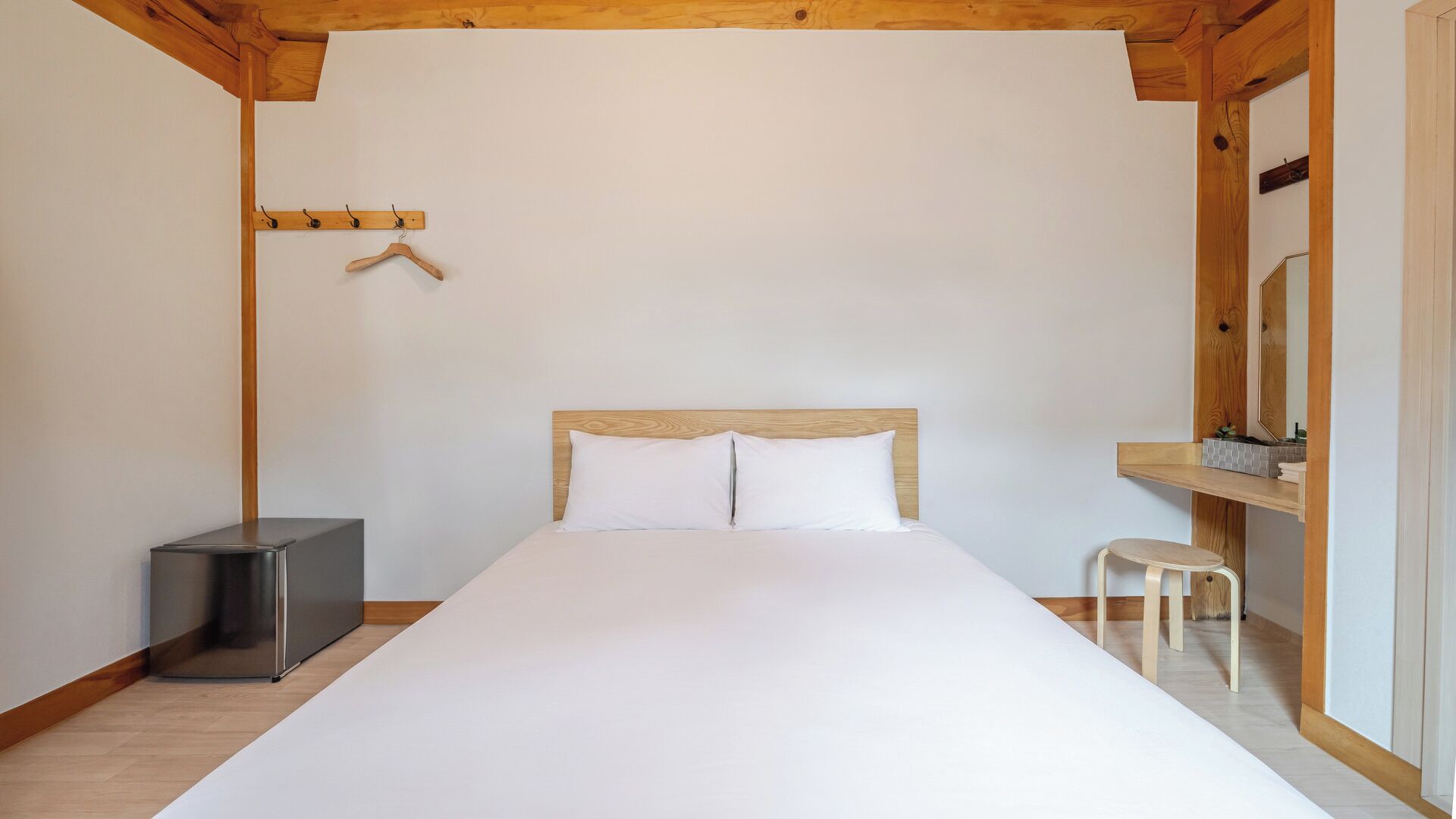 A bedroom with a bed, a wooden headboard, a hanging coat rack, a small stool, and a mirror.
