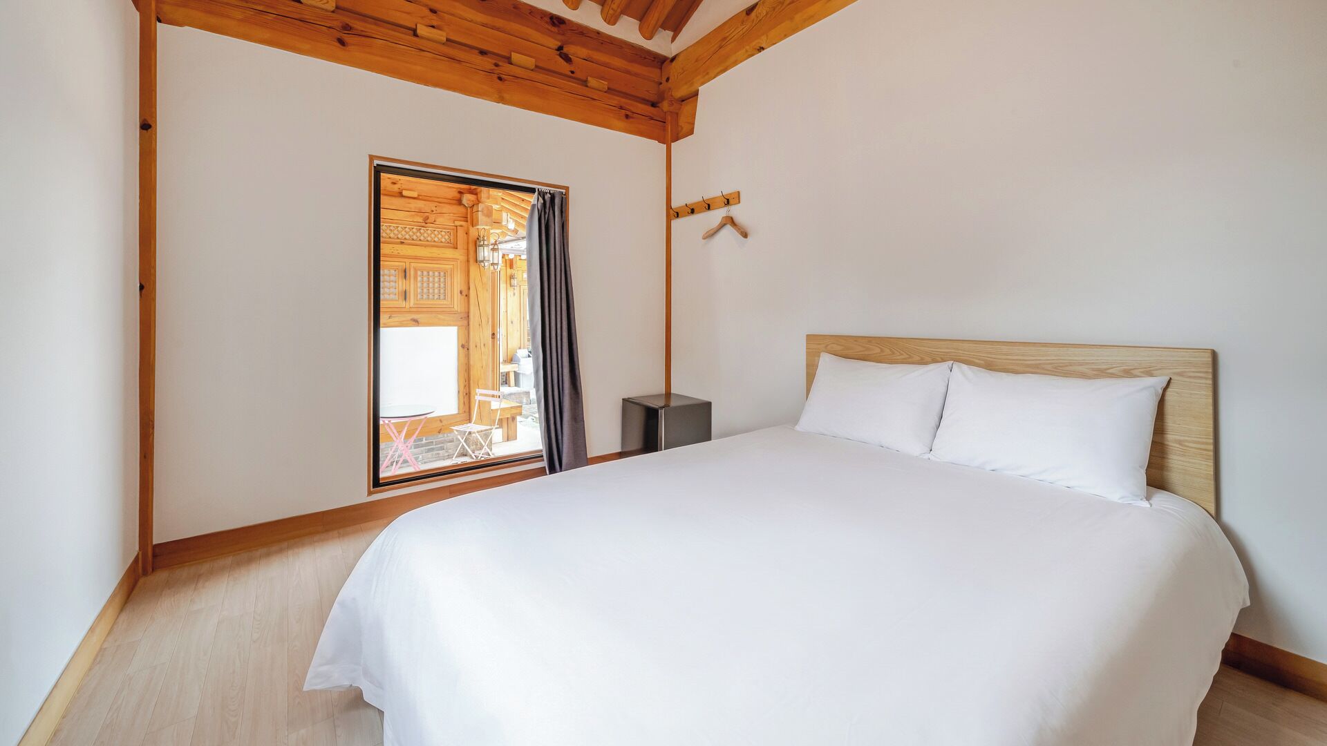 A bedroom with a wooden ceiling, a bed with white bedding, a small bedside table, and a window with a view of an outdoor area.