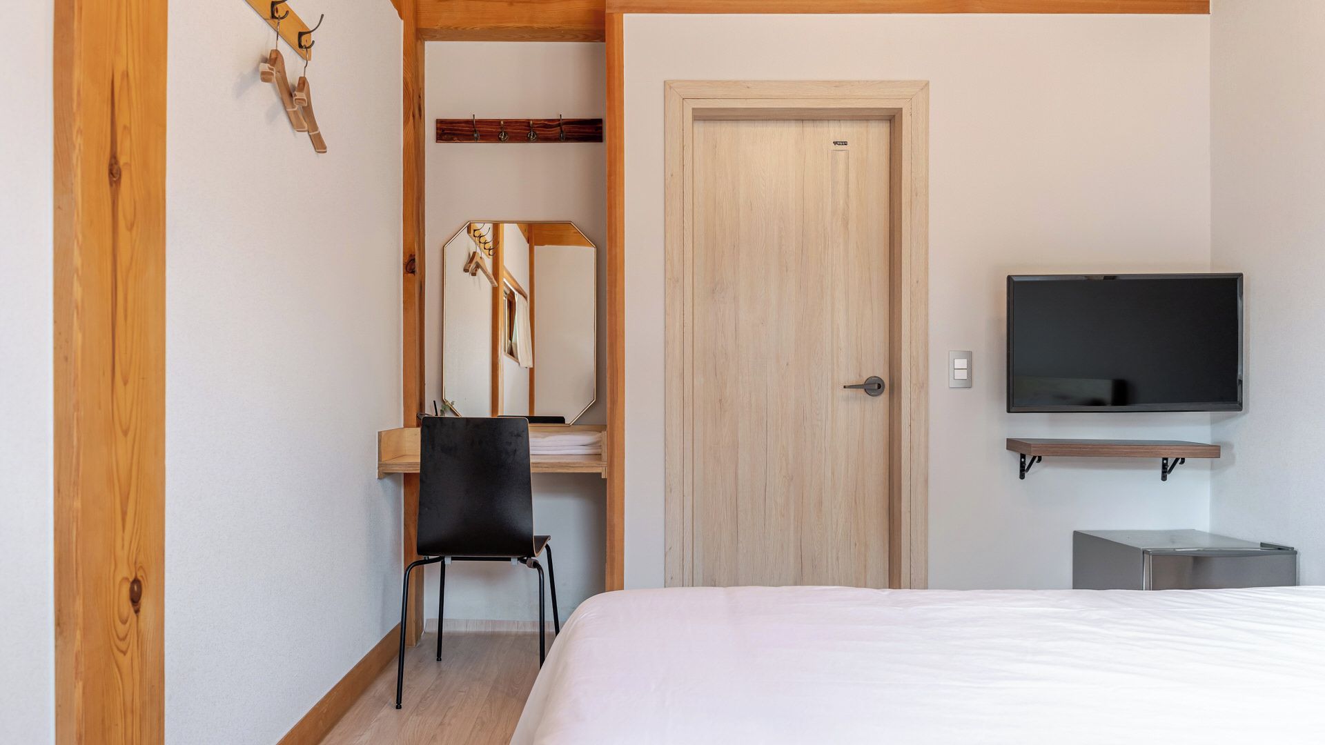 A bedroom with a bed, a desk with a chair, a television mounted on the wall, and a door.
