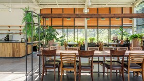 Un vaste espace intérieur aménagé en salle à manger, avec des tables et des chaises en bois, de grandes fenêtres et des plantes en pot.