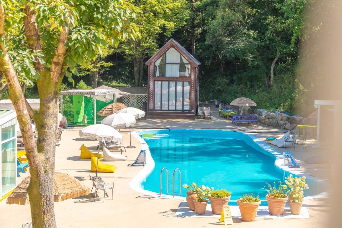 A swimming pool with lounge chairs and potted plants, surrounded by a wooden deck and a small building with a triangular roof.