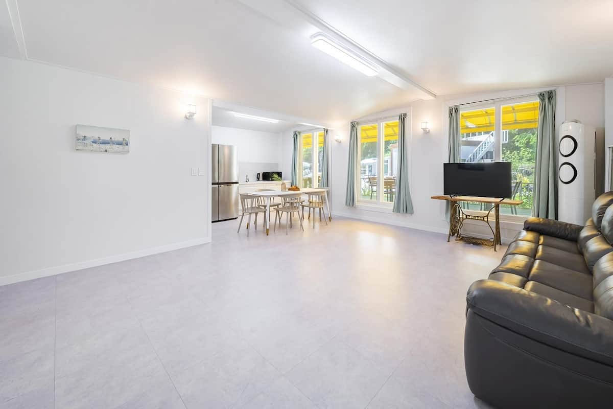 A spacious living area with a black leather sofa, a dining table with chairs, a kitchen with a refrigerator, and a TV on a stand.