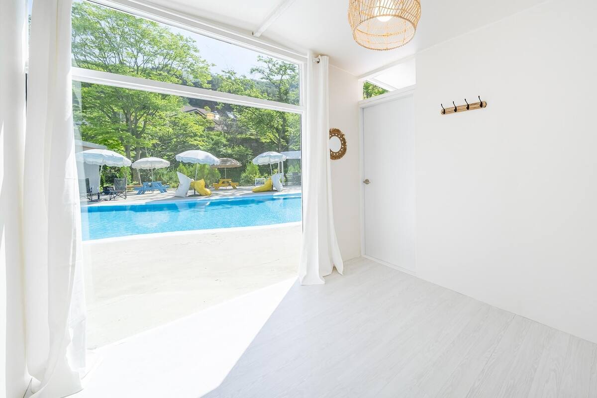 A room with a large window overlooking a swimming pool, white walls, and a wooden floor.