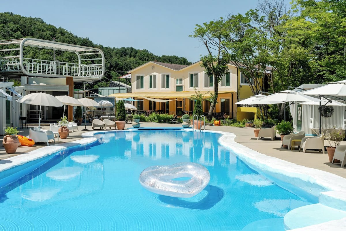 Una piscina con un flotador con forma de corazón, rodeada de mobiliario de exterior y sombrillas, con un edificio al fondo.
