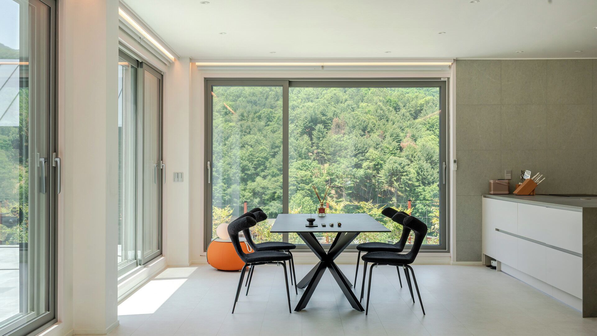 Un moderno comedor con mesa y sillas, ventanas amplias con vista al bosque y una zona de cocina.