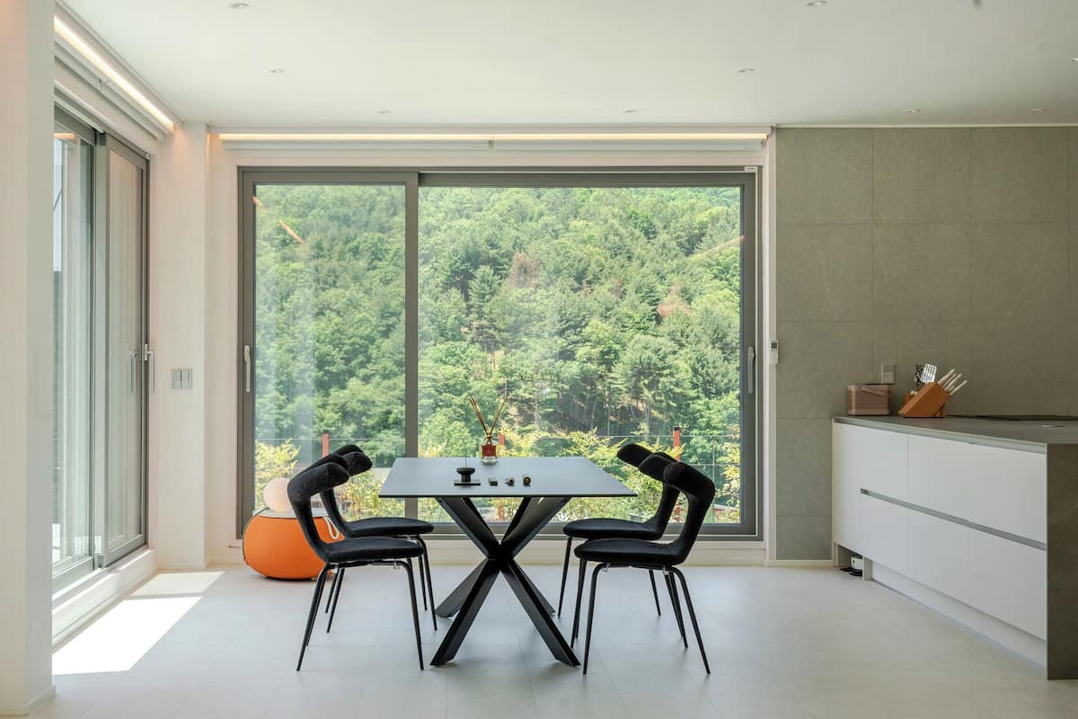 Un moderno comedor con mesa y sillas, ventanas amplias con vista al bosque y una zona de cocina.