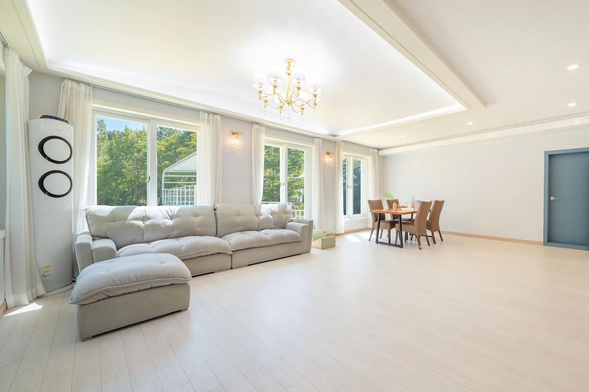 A spacious living room with a sectional sofa, a dining table with chairs, a chandelier, and a large window with curtains.