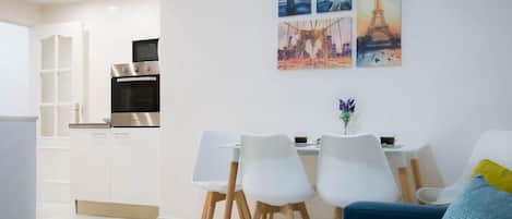 Ein modernes Wohnzimmer mit einem blauen Sofa, weißen Stühlen und einem Essbereich mit Tisch und Stühlen. Es gibt eine Küche mit Einbauofen und Kühlschrank. Die Wände sind weiß, und über dem Esstisch hängen drei Stadtansichten als Gemälde.