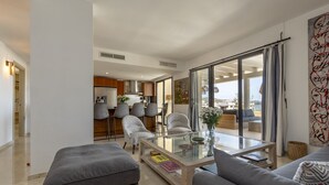 Una sala de estar moderna con una mesa de centro de vidrio, sofás grises y una zona de comedor con taburetes de bar.