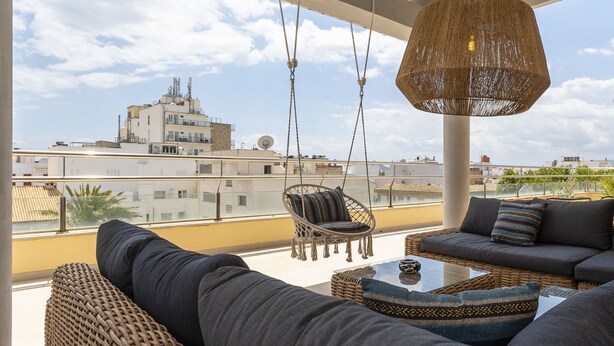 Un balcón con mobiliario de mimbre, una lámpara colgante y vistas a edificios distantes.