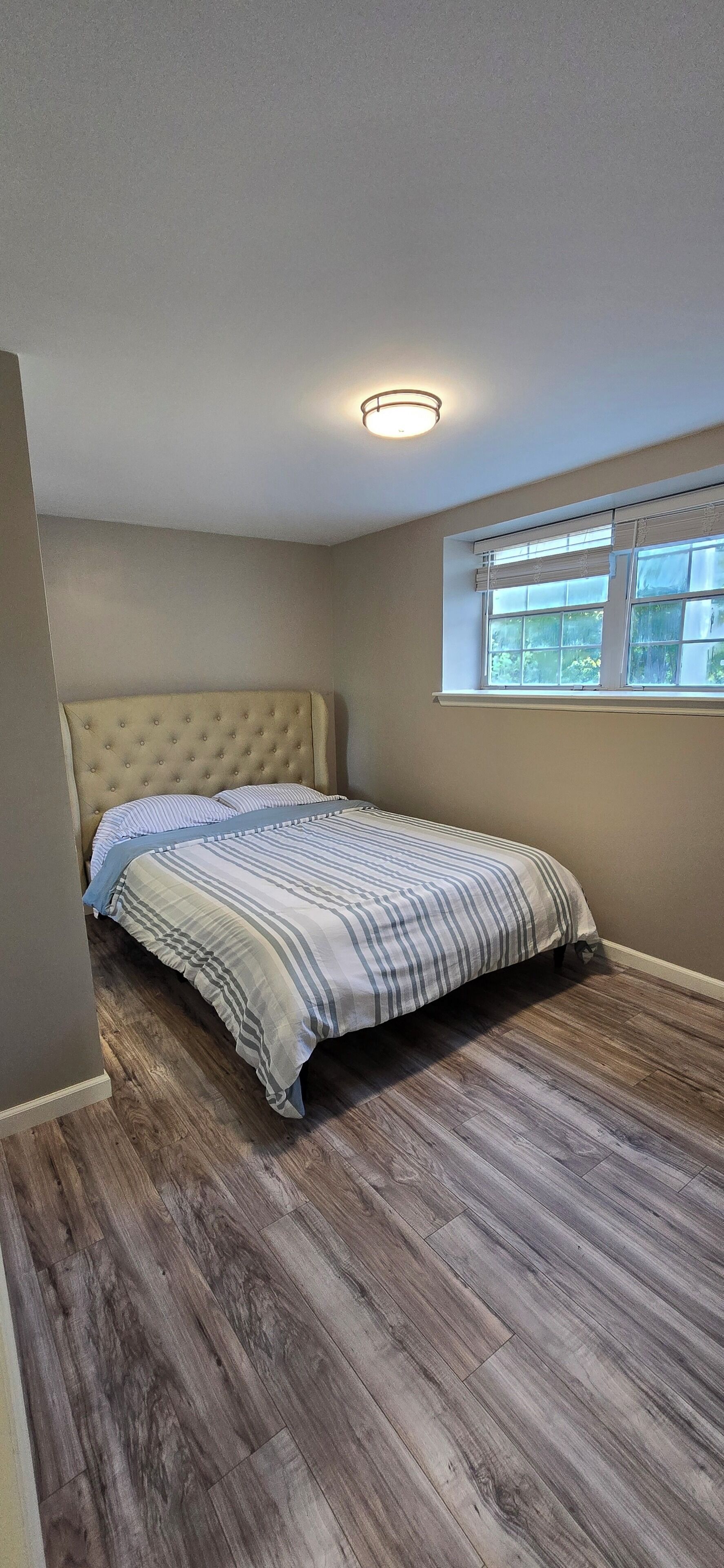 A bedroom with a tufted headboard, a bed with striped bedding, a window with blinds, and a wooden floor.