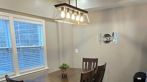 En matplats med ett träbord och stolar, en hängande lampa och ett fönster med persienner.