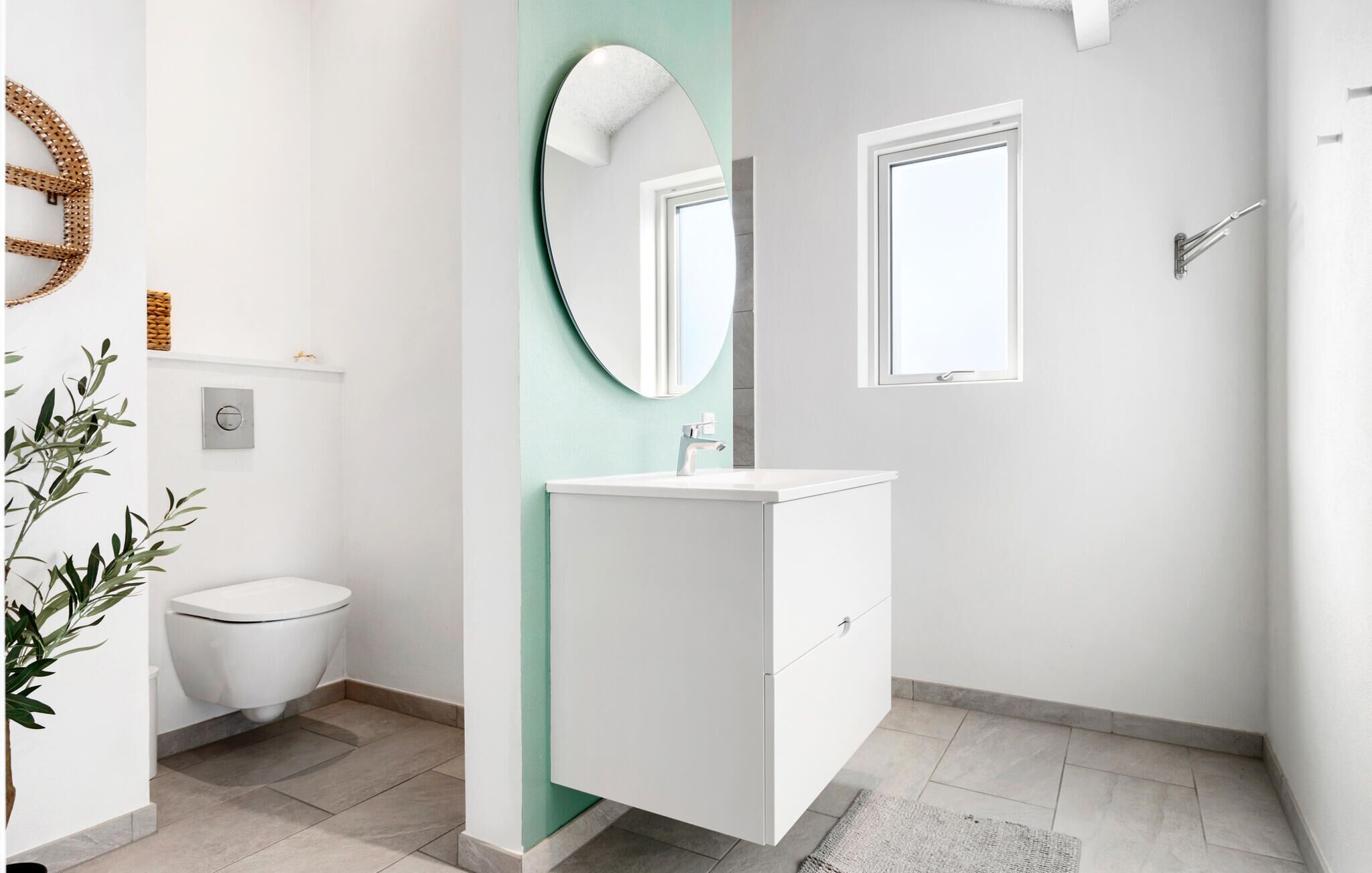 A modern bathroom with a white toilet, a white sink with a green accent wall, a round mirror, and a window.