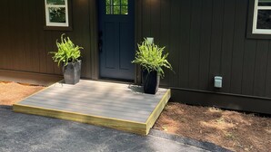 Un bâtiment à l’extérieur brun foncé, avec une porte bleue et deux plantes en pot placées sur une plateforme en bois.