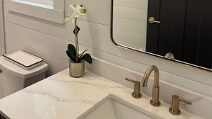 Een badkamer met een wit marmeren wastafel, gouden kranen, een zwarte deur en een raam met uitzicht op bomen.