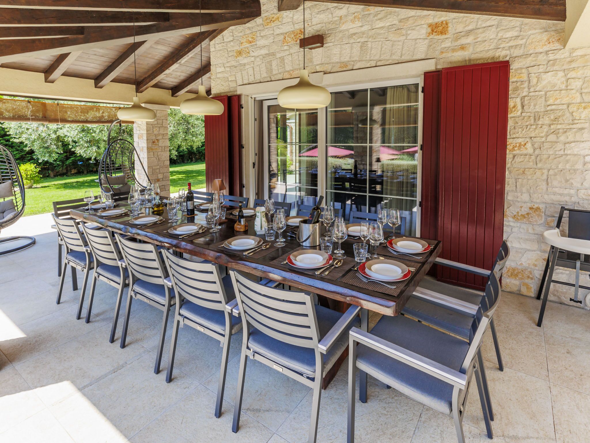An outdoor dining area with a long table set for a meal, surrounded by chairs, and a stone wall in the background.